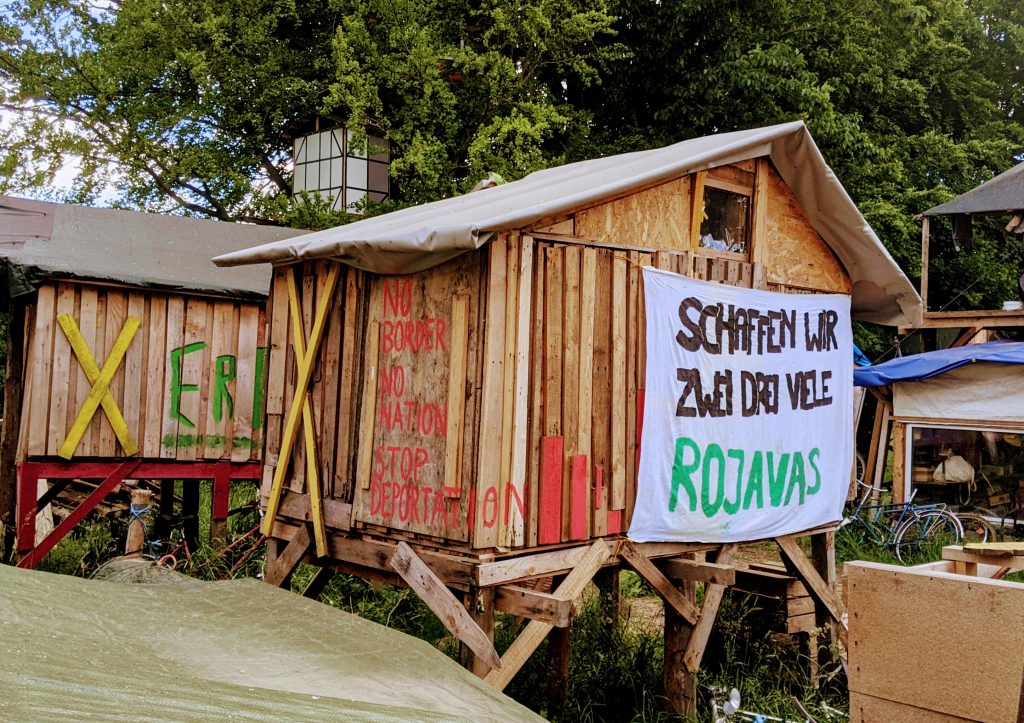 Ecological occupation to protect land from miners in Europe, with squatting structures showing a banner: "We will make two, three, many Rojavas."