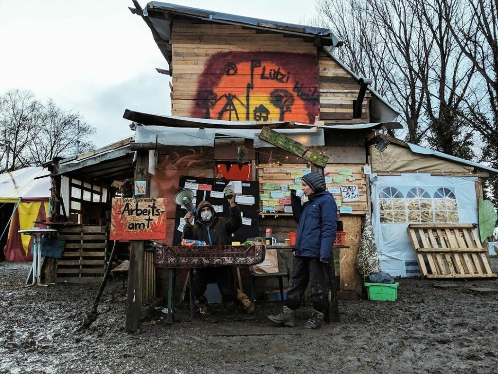 A two-story shack structure made of wood, painted with street art, with billboards in front.  A masked activist is posing by a stand in the front like an announcer, next to the  sign which reads: "Burning job center".