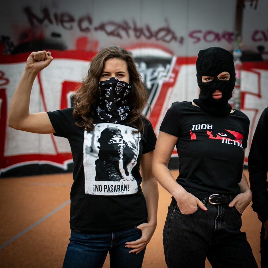 A photo showing two female-presenting people.  One is wearing a black bandana with the trans-feminism symbol and a black T-shirt featuring a masked antifa, labelled with the slogan "¡No Pasarán!".  She's posing with one fist raised.  The other one wears a black, three-hole balaclava and a black T-shirt showing an intense stare in red, with the brand logo for Mob Action.  She's posing with hands in nice black denim pants and a stare as intense as the T-shit logo.